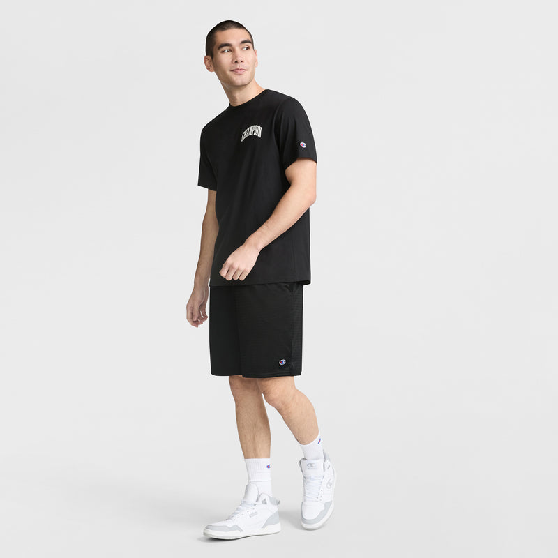 A person wearing a black Champion t-shirt, Champion 9" Classic Mesh Shorts with C Logo, white crew socks, and white sneakers stands against a plain white background looking to the side with one arm slightly raised.