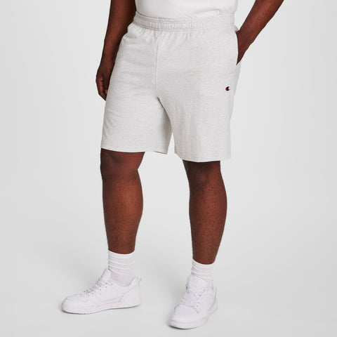A person wearing Champion 9" Lightweight Lounge Shorts, C Patch Logo - Big & Tall in light gray, a white T-shirt, white socks, and white sneakers stands against a plain light background.