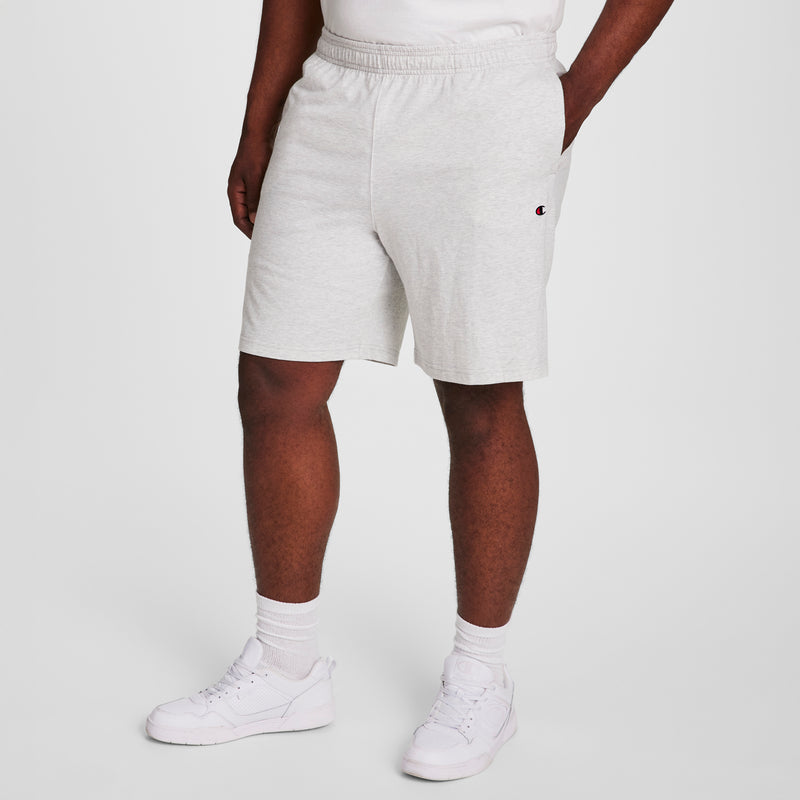 A person wearing Champion 9" Lightweight Lounge Shorts, C Patch Logo - Big & Tall in light gray, a white T-shirt, white socks, and white sneakers stands against a plain light background.
