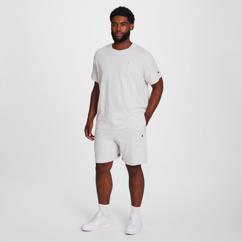 A man with short curly hair and a beard stands against a plain white background, wearing Champion 9" Lightweight Lounge Shorts with C Patch Logo (Big & Tall), a light gray t-shirt, white socks, and sneakers, hand at his side.