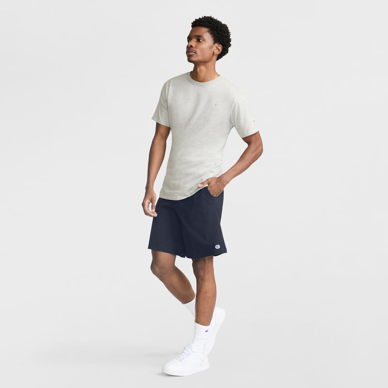 A man stands looking to the side wearing a light gray T-shirt, Champion 9" Lightweight Lounge Shorts with C Patch Logo in navy, white crew socks, and white sneakers against a plain light background.