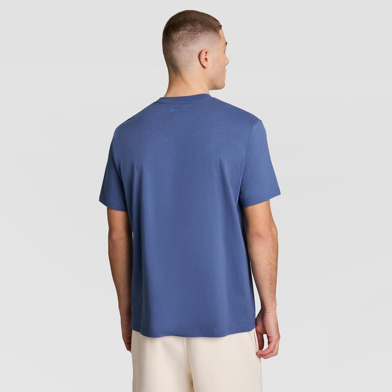 A man with short hair faces away, wearing a Champion Classic Graphic T-Shirt with a tonal script logo and light-colored shorts against a plain background.