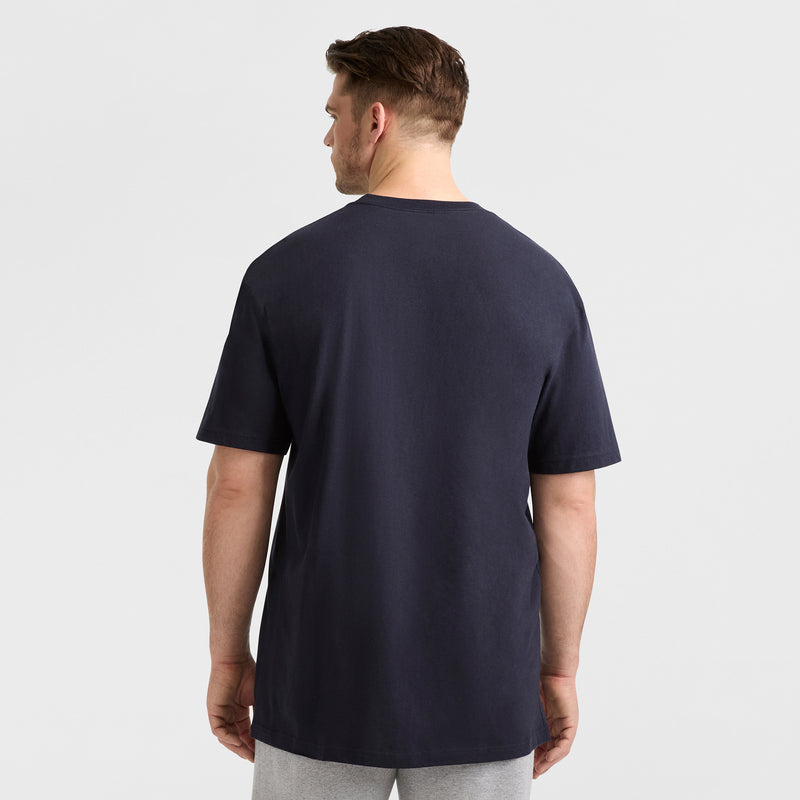 A man with short brown hair stands with his back to the camera, wearing a Champion Classic Graphic T-Shirt, Script Logo - Big & Tall in navy blue and light gray pants against a plain white background.