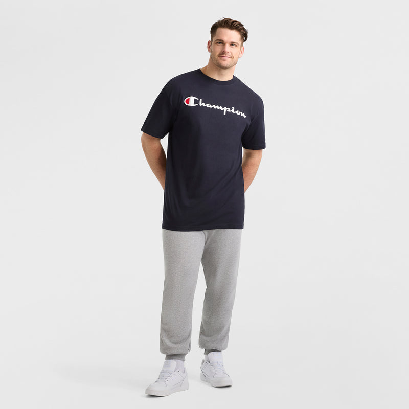 A man stands against a plain white background wearing the Champion Classic Graphic T-Shirt, Script Logo - Big & Tall in navy blue, gray sweatpants, and white sneakers—smiling with his hands behind his back.