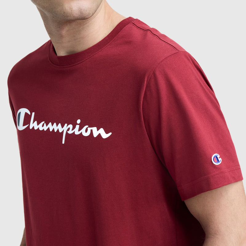 A person wears the Champion Classic Graphic T-Shirt, Script Logo—a men’s streetwear staple. The white Champion script logo is across the chest with a small embroidered emblem on the left sleeve. Background is plain light gray.