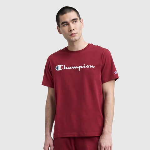 A young man with short dark hair wears a Champion Classic Graphic T-Shirt featuring the Script Logo and matching red shorts, showcasing a streetwear vibe against a plain light gray background.