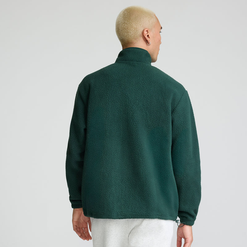 A person with short blond hair is shown from the back, standing against a plain light background, wearing a dark green Champion Micro Fleece 1/4 Zip and light gray pants.