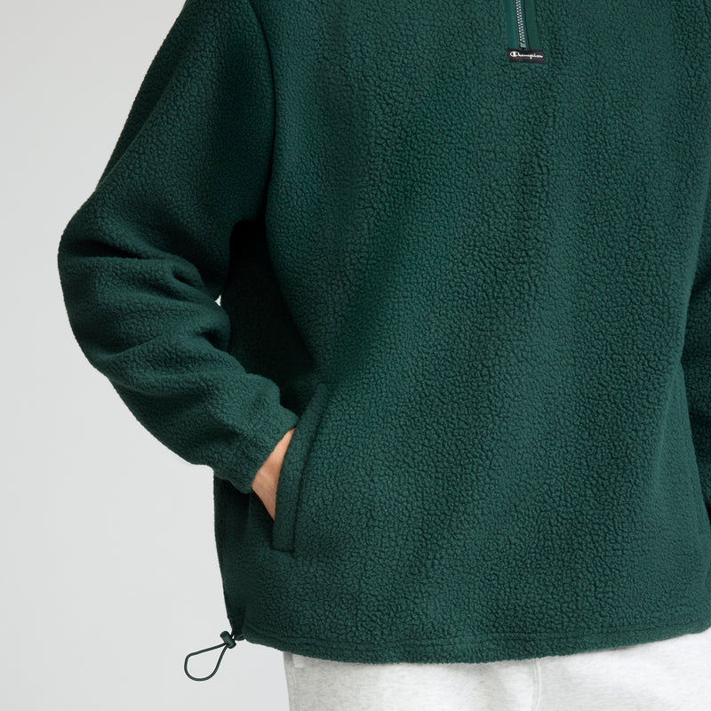 A person wears a Champion Micro Fleece 1/4 Zip in dark green with hands in pockets; only the torso and part of the arms show, paired with light gray pants.