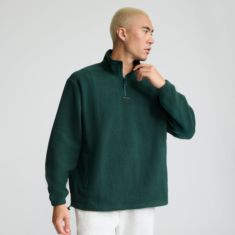 A man with short bleached hair wears the Champion Micro Fleece 1/4 Zip in dark green and light gray pants, standing against a plain light background and looking to his left.