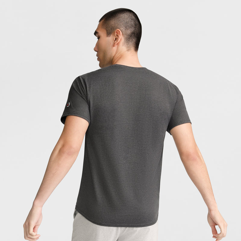 A man with short dark hair stands with his back to the camera, wearing a Champion Classic T-Shirt, C Logo in dark gray and light pants against a plain light background.