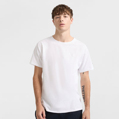 A young man with short brown hair faces forward, showing his left forearm tattoo while wearing the Champion Classic T-Shirt, C Logo, in a soft cotton blend and dark pants against a plain light gray background.