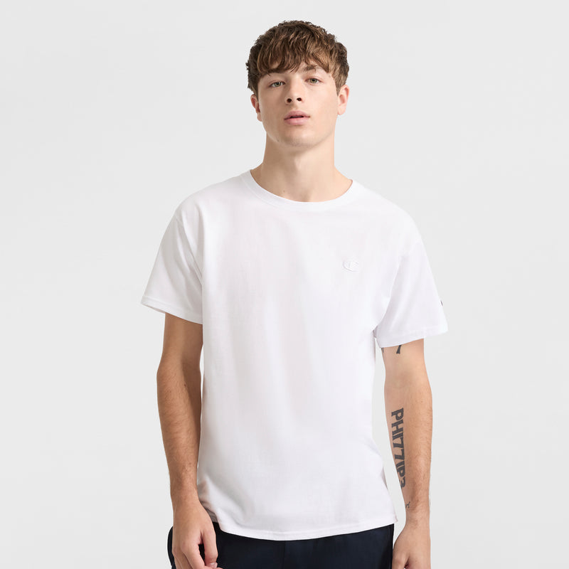 A young man with short brown hair faces forward, showing his left forearm tattoo while wearing the Champion Classic T-Shirt, C Logo, in a soft cotton blend and dark pants against a plain light gray background.