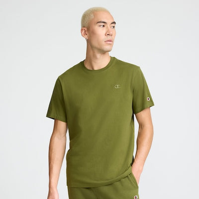 A man with short bleached blond hair wears an olive green T-shirt and matching pants. He looks to his left with one hand in his pocket, standing against a plain light gray background.