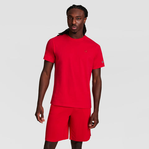 A man with long braided hair wears a matching red Champion Classic T-Shirt with C Logo and red shorts, standing against a plain light gray background.