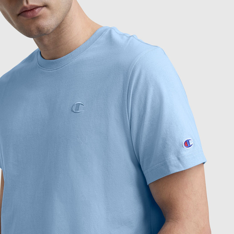A man wears a light blue Champion Classic T-Shirt, C Logo, with the embroidered logo on the chest and sleeve. Only his upper body and lower half of his face are visible against a plain light gray background.