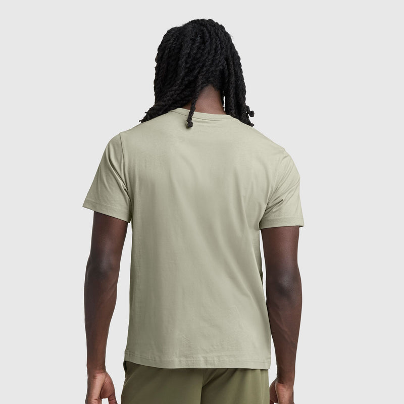 A person with long, twisted hair stands with their back to the camera, wearing a light olive green Champion Classic T-Shirt, C Logo, and matching shorts against a plain gray background.