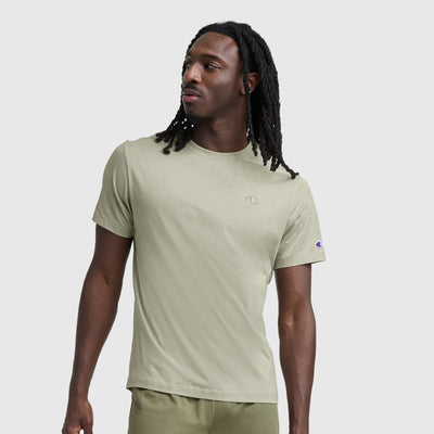 A man with long dreadlocks stands against a plain light gray background, wearing the Champion Classic T-Shirt, C Logo, and matching shorts, looking off to the side.