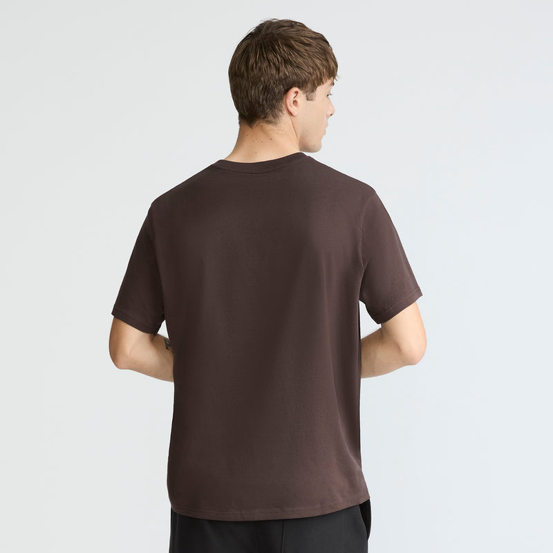 A man with short brown hair stands with his back to the camera, wearing a Champion Classic T-Shirt, C Logo in brown and black pants against a light gray background.