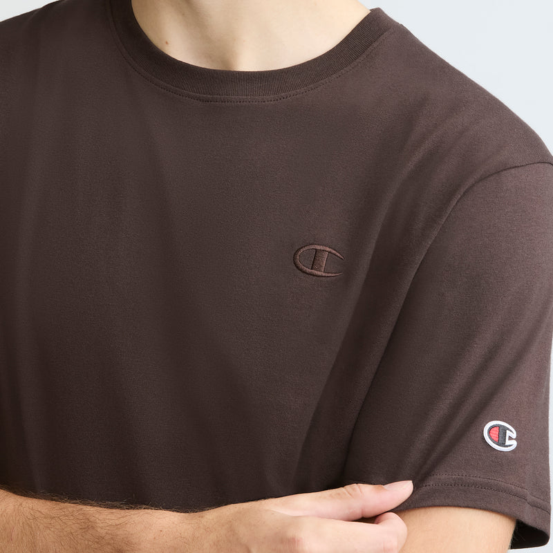 A person with arms crossed, shown from the shoulders down, wears a brown Champion Classic T-Shirt, C Logo, featuring a crewneck and logo on the chest and sleeve.