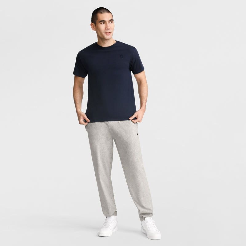 A person stands against a plain background wearing a navy blue Champion Classic T-Shirt with C Logo, light gray sweatpants, and white sneakers, with one hand in their pocket and the other by their side.