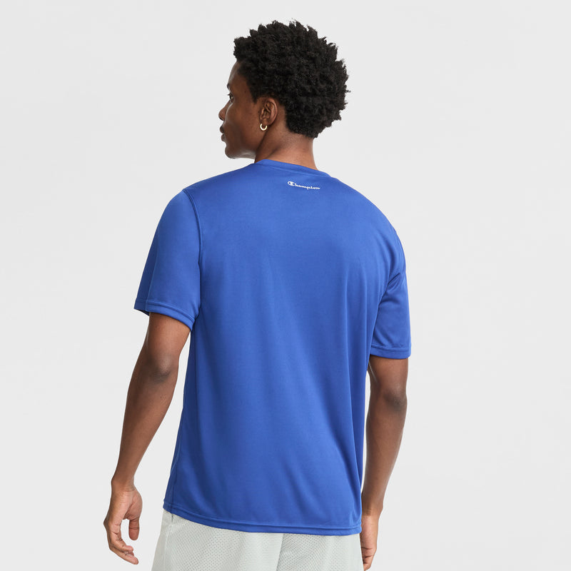 A person faces away from the camera in a Champion Sport T-Shirt, Reflective C Logo. The royal blue men’s tee features Double Dry tech for an athletic vibe, with light shorts and a small white logo at the neckline against a plain backdrop.