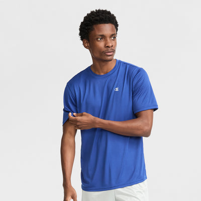 A man in a royal blue Champion Sport T-Shirt with a reflective C logo and light shorts stands against a white background, looking left while adjusting his sleeve.