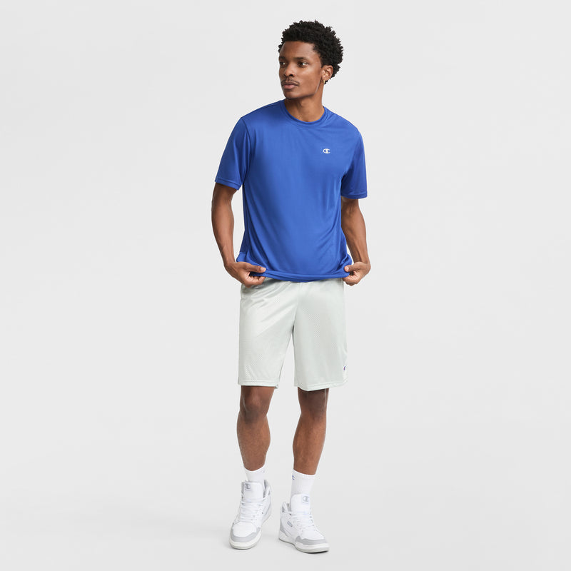 A man poses against a white background, wearing a blue Champion Sport T-Shirt with Reflective C Logo and Double Dry tech, paired with light gray shorts, white socks, and sneakers. He looks to the side, holding his shirt for an athletic vibe.