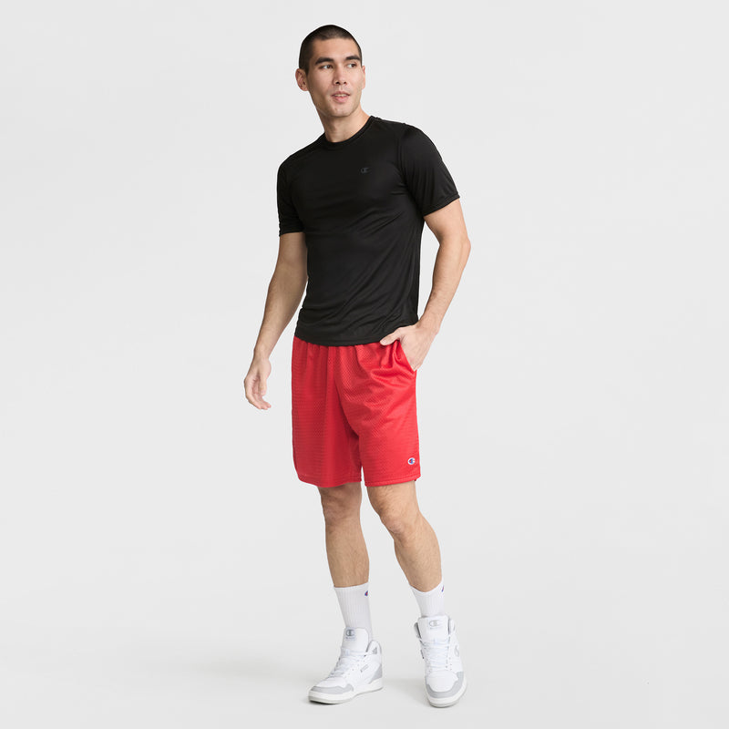 A man stands against a white background wearing a Champion Sport T-Shirt with Reflective C Logo, red athletic shorts, white crew socks, and white sneakers. He has one hand in his pocket and looks slightly to the side.