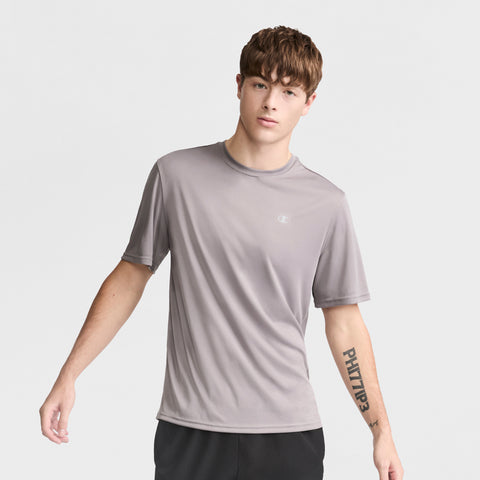 A young man with short brown hair models the Champion Sport T-Shirt with Reflective C Logo and black shorts, standing against a white background. His left arm, featuring a forearm tattoo, is slightly extended.