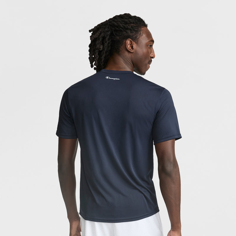A man with braided hair is seen from the back wearing a navy Champion Sport T-Shirt with a Reflective C Logo and white shorts, set against a plain white background.