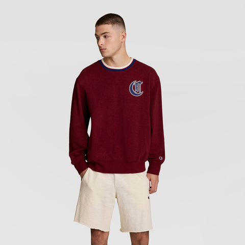 A young man wears the Champion Modern Retro Washed Crew Sweatshirt with a C logo and beige shorts, standing with hands in pockets and looking to the side against a plain light background.