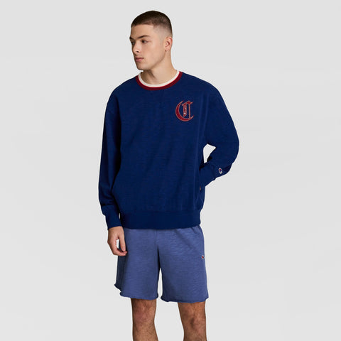 A person stands against a plain background in a blue Champion Modern Retro Washed Crew Sweatshirt, featuring a heavy textured slub cotton and red and white C Logo, paired with blue athletic shorts for vintage streetwear style.
