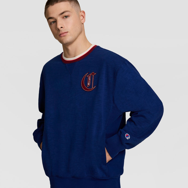 A young man stands hands-in-pockets against a plain background, sporting the Champion Modern Retro Washed Crew Sweatshirt with a large embroidered C logo and red-and-white collar, channeling vintage streetwear style.