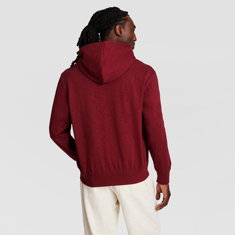 A person with long, dark, braided hair is seen from the back wearing a Champion Modern Retro Washed Hoodie with a C Logo and light-colored pants, standing against a plain light gray background.