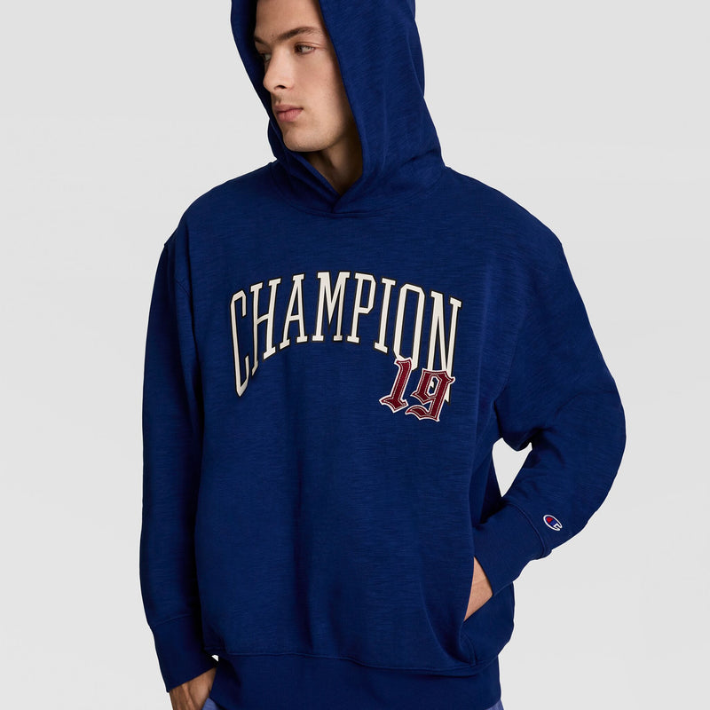 A person in a blue Champion Modern Retro Washed Hoodie with a bold C logo on the front looks to the side, one hand in their pocket.