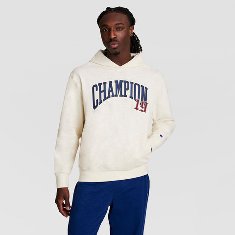 A man wears a cream-colored Champion Modern Retro Washed Hoodie with a C logo, paired with blue sweatpants, standing against a plain light gray background.