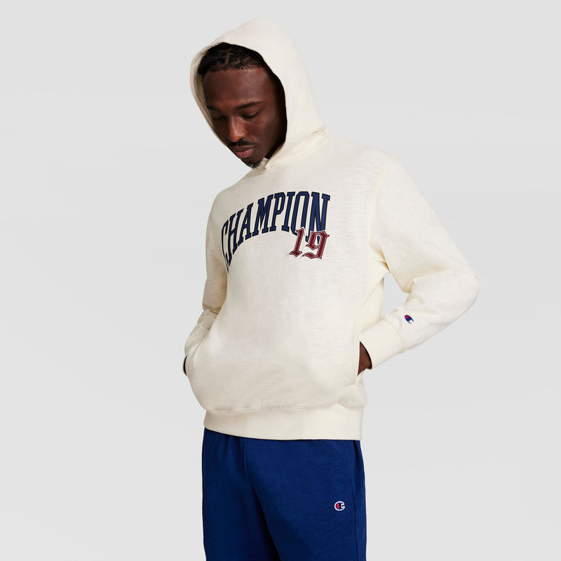 A man wears a Champion Modern Retro Washed Hoodie with C Logo and blue sweatpants, standing with hands in pockets and looking down against a plain light gray background.