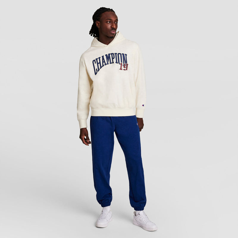 A man stands against a plain background wearing a cream Champion Modern Retro Washed Hoodie with the C logo, blue sweatpants, and white sneakers. He looks to his left with one hand by his side.