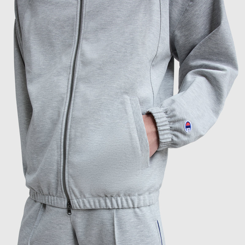 A person wears a light gray Champion Gameday Full Zip Mock Neck tracksuit, featuring elastic cuffs and waistband, with their right hand in the jacket pocket. A small embroidered C Logo appears on the sleeve.
