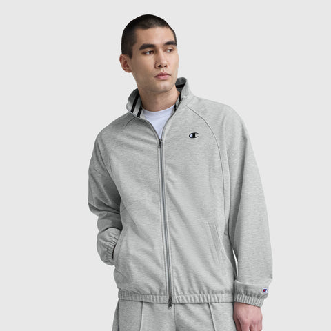 A man stands with one hand in his pocket against a plain light background, wearing a light gray Champion Gameday Full Zip Mock Neck jacket featuring an embroidered C logo and matching pants.