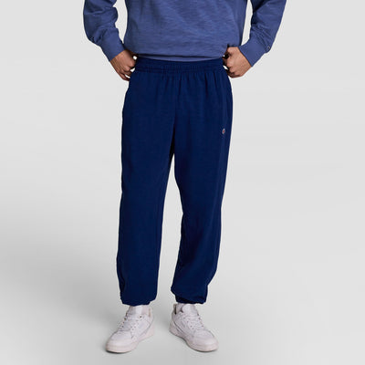 A person stands hands-on-hips in Champion’s Modern Retro Washed Pant with C Logo and a blue Champion sweatshirt. White sneakers finish the look. The face is not visible, and the background is plain and light.