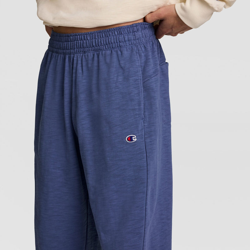 A person wears Champion’s Modern Retro Washed Pant, C Logo in blue with the signature embroidery, paired with a light sweatshirt. Only their waist and upper legs are visible as one hand rests on their hip.