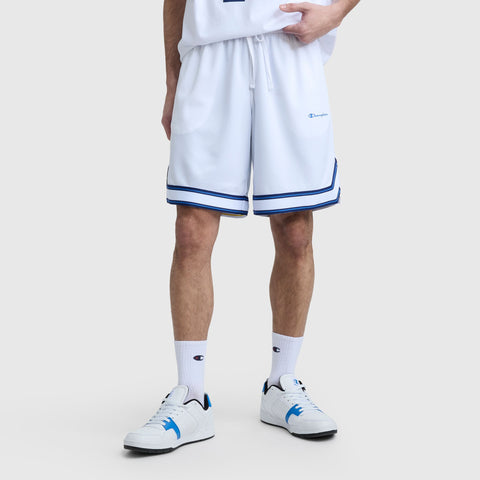 Someone stands against a light gray background wearing Champion's 10" Gridiron Reversible Short with blue trim, a matching white shirt, white crew socks, and white sneakers featuring blue accents.