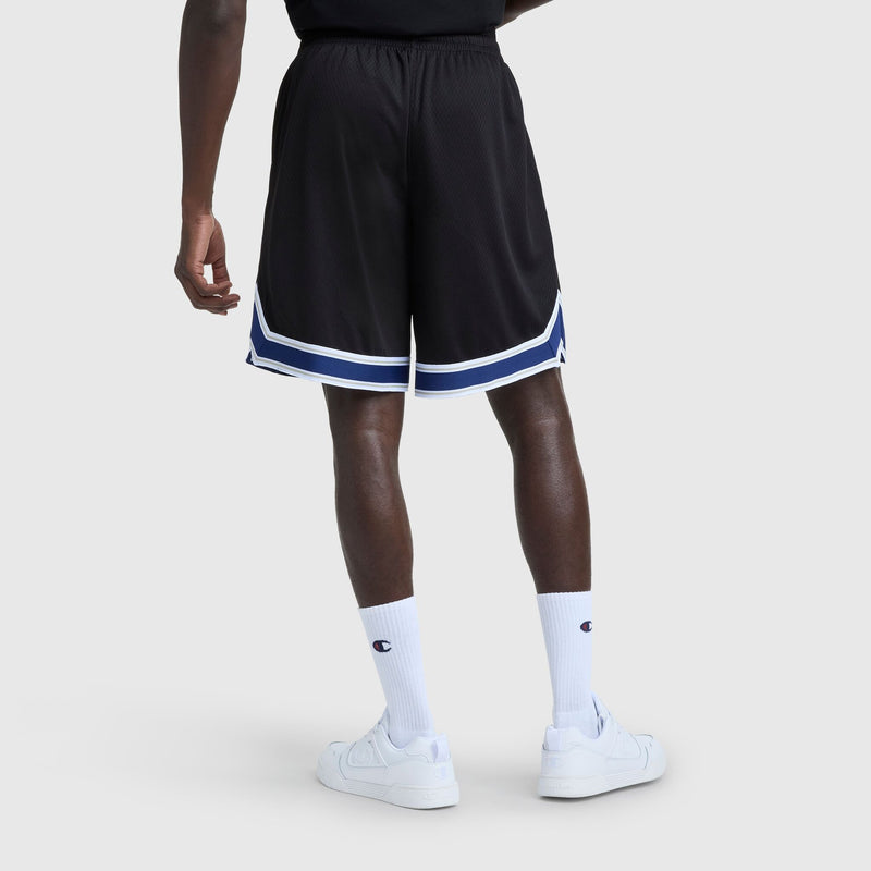 A person wearing black Champion 10" Gridiron Reversible Short with script logo, featuring blue and white stripes, paired with white crew socks displaying a logo and white sneakers, stands against a plain light gray background.