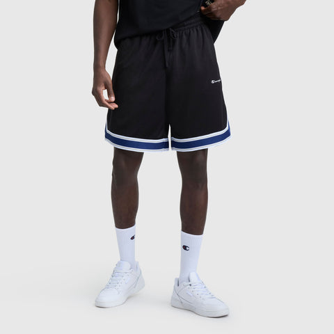 A person wearing black Champion 10" Gridiron Reversible Shorts with Script Logo and white and blue trim, a black shirt, white crew socks with a small logo, and white sneakers stands against a plain light gray background.