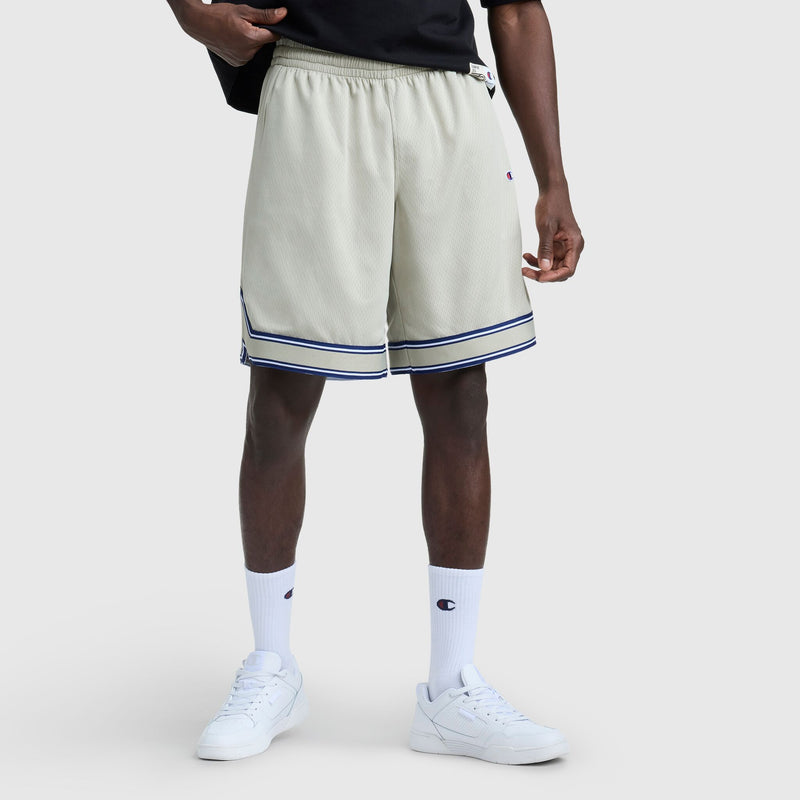 Someone stands against a plain light gray background wearing cream-colored Champion 10" Gridiron Reversible Shorts with a script logo and blue stripes, a black shirt, white socks, and white sneakers.