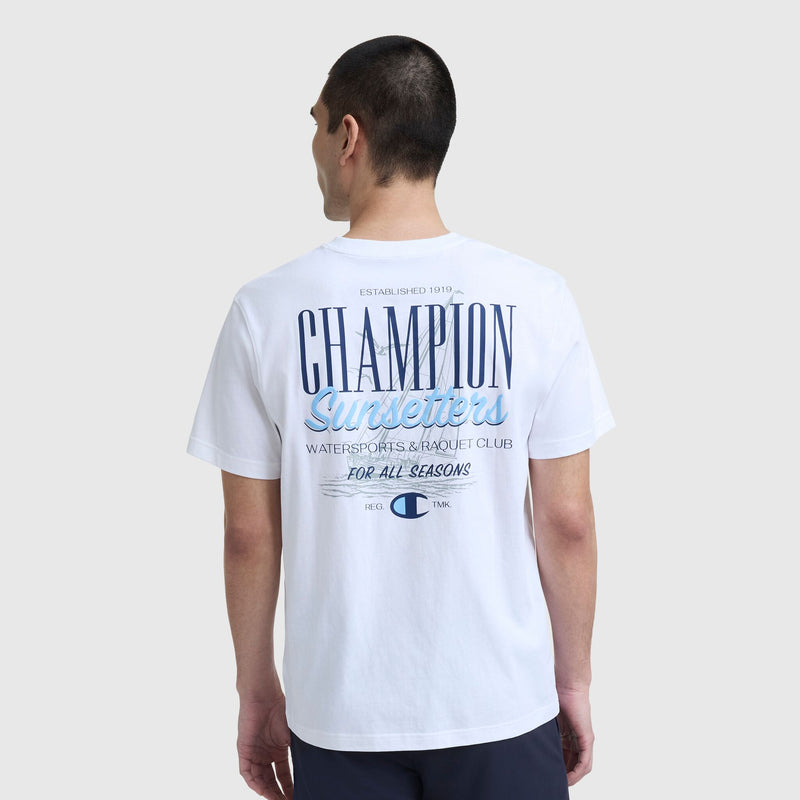 A man with short dark hair, seen from behind, wears the Champion Sunsetters Graphic T-Shirt, C Logo—a men's cotton tee with blue CHAMPION Surfers Watersports & Racquet Club For All Seasons text and small logos.