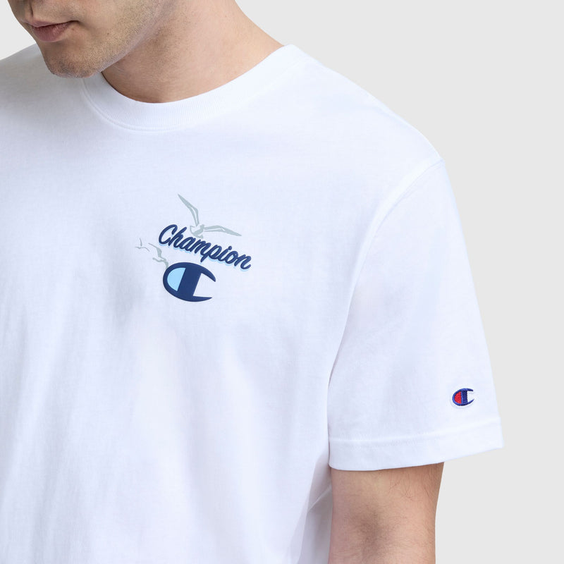 A man wears a white Champion Sunsetters Graphic T-Shirt, featuring the C logo and a small bird graphic on the chest, plus a Champion patch on the left sleeve. The men's cotton tee offers casual comfort and style against a plain light background.