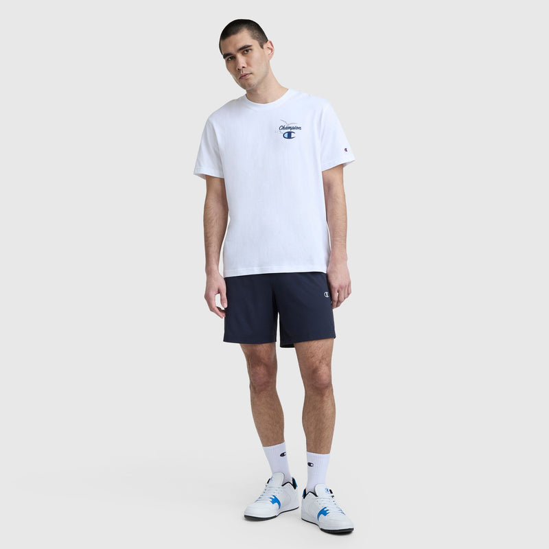 A man with short dark hair and a neutral expression stands against a plain background, wearing a white Champion Sunsetters Graphic T-Shirt with C Logo, navy blue shorts, white crew socks, and white sneakers with blue accents.
