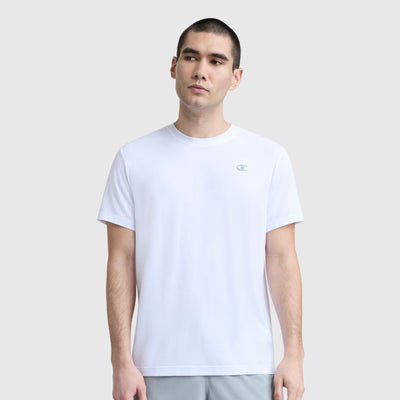 A man with short dark hair wears a Champion Sport Crewneck T-Shirt, Script Logo—a plain white men's slim-fit tee—paired with light gray shorts. He stands against a light gray background, looking slightly to the side.